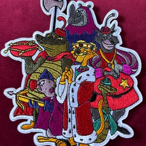 May include: Embroidered patch featuring a group of cartoon characters, including a crocodile, rhino, and fox, all wearing regal attire. The patch has a white border and is set against a burgundy background. The characters are in various colors, including green, purple, and red.
