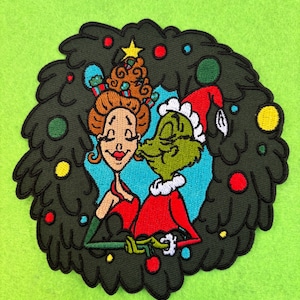 May include: An embroidered patch featuring the Grinch and Cindy Lou Who in a Christmas wreath. The Grinch is wearing a Santa hat and Cindy Lou Who is wearing a red dress and a green bow in her hair.