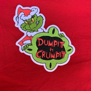 May include: Green and white embroidered patch featuring the Grinch from the cartoon "How the Grinch Stole Christmas" wearing a Santa hat. The Grinch is holding a green sign that says "Dump It To Crumpit".