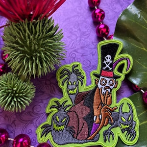 May include: An embroidered iron-on patch featuring a green-skinned character with a top hat and a skull and crossbones design. The character is surrounded by four green, glowing, ghost-like figures. The patch is on a purple background.
