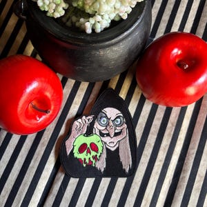 May include: A black patch featuring a cartoon witch holding a green skull-shaped apple. Two red apples and a black cauldron with white flowers are also visible. The items are arranged on a black and white striped surface.