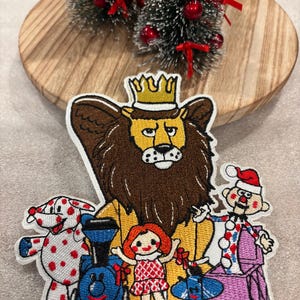 May include: Embroidered patch featuring a lion wearing a crown, surrounded by toy characters including a dog, a train, a doll, a plane, and a clown. The patch has a white border and is set against a wooden background with miniature Christmas trees.