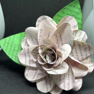 May include: A delicate paper flower crafted from the pages of a book. The flower is a light pink color with a green leaf. The flower is made up of several layers of paper, each with text printed on it. The text is visible and includes phrases like "earth crack", "well anymore", "He would have", "the entire", "Awen", "and", "Mel with", "run", "you, Jak", "a glance", "orgive", "v", "bow across", "a viol", "warn", "all."
