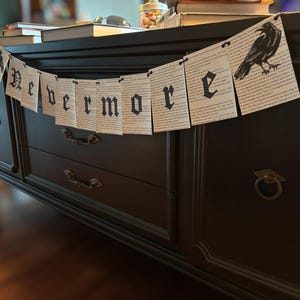 May include: A black banner with the word "Nevermore" in gothic lettering and a raven illustration. The banner is made of paper and is draped over a dark wooden cabinet. Books and other items are on top of the cabinet.