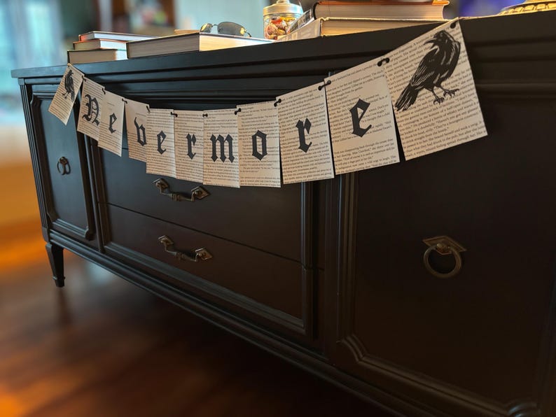 May include: A black banner with the word "Nevermore" in gothic lettering and a raven illustration. The banner is made of paper and is draped over a dark wooden cabinet. Books and other items are on top of the cabinet.