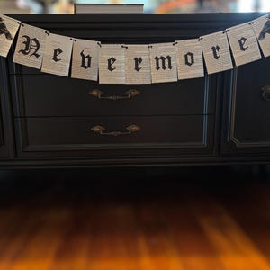 May include: A black banner with the word "Nevermore" in gothic lettering hangs above a black dresser. The banner is made of paper with printed text and features a raven illustration on each end. The dresser has two drawers and decorative hardware.