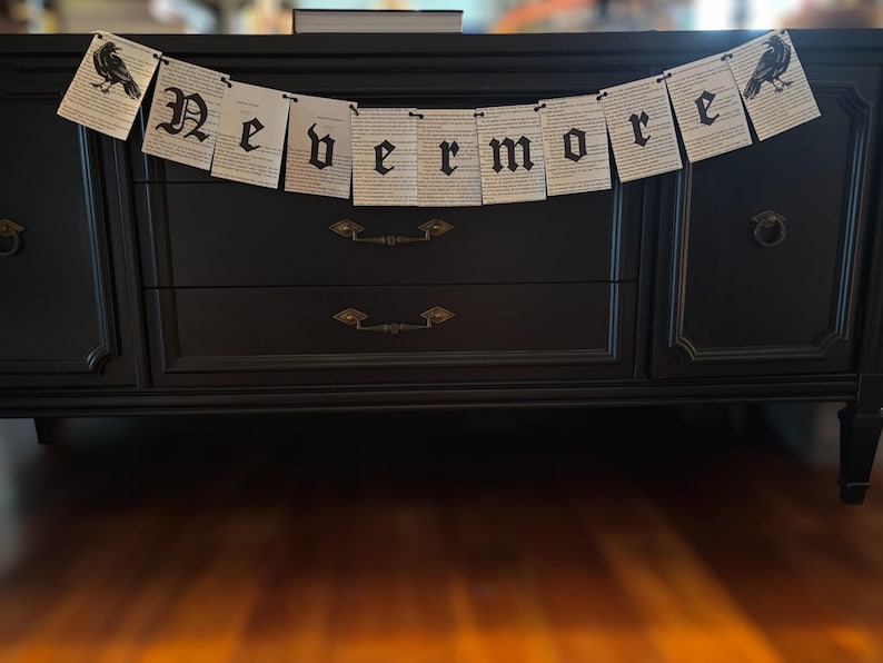 May include: A black banner with the word "Nevermore" in gothic lettering hangs above a black dresser. The banner is made of paper with printed text and features a raven illustration on each end. The dresser has two drawers and decorative hardware.