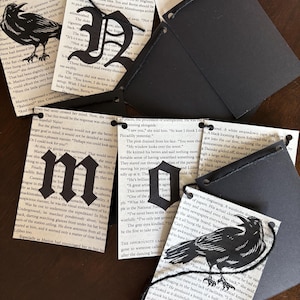 May include: Black paper tags with cut-out letters and raven silhouettes. The tags are attached to pages from a vintage book. The letters spell out words and phrases. The tags are connected with black string.