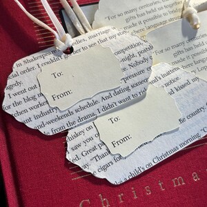 May include: Close-up of gift tags with torn edges, attached to a red book with gold lettering. The tags are cream-colored with the words "To:" and "From:" printed on them. White ribbons are attached to the tags.