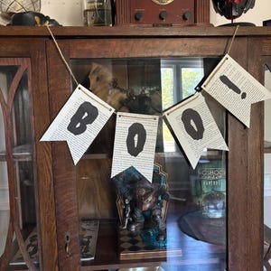 May include: A wooden cabinet with glass doors displays a collection of items. A banner spelling "BOO!" hangs in front. The banner is made of book pages with black letters. Other items include a vintage radio, perfume bottles, and a decorative figure.