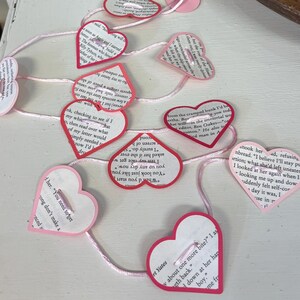 May include: A handmade garland of heart-shaped paper cutouts with text from a book, strung together with pink thread. Each heart is outlined in red and has a button attached. A charming, romantic decoration.