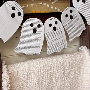 May include: A Halloween-themed decoration featuring paper ghost cutouts with black eye and mouth details, strung on a twine garland. The ghosts are made from book pages. A cream-colored textured blanket is draped below.