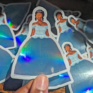 May include: A stack of iridescent blue princess stickers with a black woman wearing a blue dress and a crown. The stickers have a holographic finish and the text "@rantuknoteembroidery" is visible on one of the stickers.