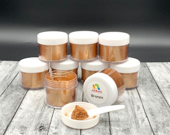May include: Eight small jars of bronze colored mica powder, with one jar open and a small scoop of powder in the lid. The jar has a label that says "Aroma  Essentials  Bronze".