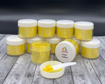 May include: A set of ten small jars filled with yellow pearl pigment powder. The jars have white lids and are arranged in a pyramid shape. A small white scoop is filled with the powder and is sitting next to the jars. The label on one of the jars reads "Aroma Effects Pearl Yellow".
