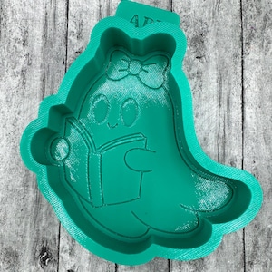 May include: A teal-colored cookie cutter in the shape of a cartoon ghost reading a book. The ghost has a bow on its head and a smiling face. The cutter has the letters "AP" on the top.