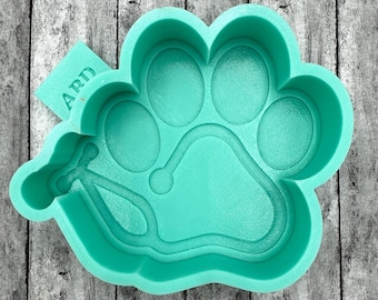 May include: A teal blue silicone mould shaped like a paw print with a stethoscope design inside. The mould is on a wooden surface. The word "ARD" is visible on a small tag.