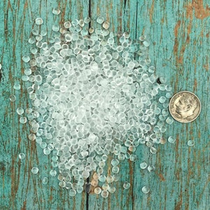 May include: A large pile of clear, translucent, round beads on a blue and green wooden surface. The beads are small and uniform in size.