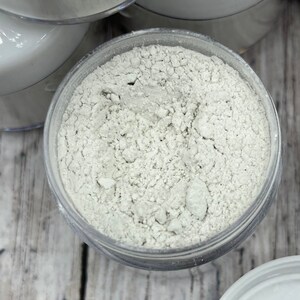 May include: A close-up view of a clear plastic container filled with a white powder. The powder is finely ground and has a slightly grainy texture.