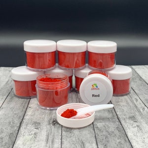 May include: A set of seven small clear jars filled with red food coloring powder. The jars have white lids and are stacked in a pyramid shape. The jar at the bottom of the pyramid has a white label with the text "Aroma Food Colors" and "Red".