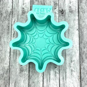 May include: A teal blue silicone mold shaped like a spider web. The mold has a raised design of a spider web and says "ARD" on the side.