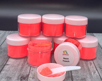 Puede incluir: Un grupo de ocho pequeños frascos de pigmento en polvo de color melocotón neón. Los frascos están apilados en forma de pirámide, con un frasco abierto y una pequeña pala blanca en su interior. La etiqueta del frasco abierto dice "Aroma Cake Art, Neon Peach."