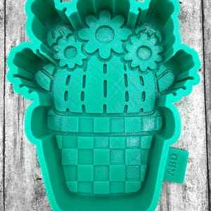 May include: Green silicone mold shaped like a cactus in a pot with flowers. The pot has a checkered pattern. The mold is labeled "ABD".