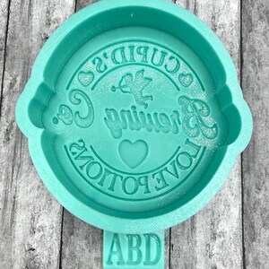 May include: A teal green silicone mold with the text "Cupid's Brewing Co. Love Potions" and a small heart in the center. The mold has a small rectangular tag with the letters "ABD" on it.