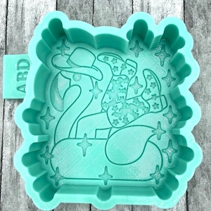 May include: A light green silicone mold for making a cake or cookie shaped like a person wearing a cowboy hat and boots riding a flamingo float. The mold has a textured surface and is surrounded by a decorative border.