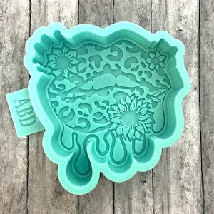 May include: A light green silicone mold shaped like a pair of lips with a leopard print design and sunflowers. The mold is dripping with a liquid effect.
