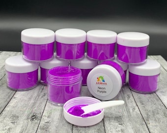 Puede incluir: Varios frascos pequeños de polvo de pigmento morado neón, con un frasco abierto y una pequeña pala blanca en su interior. Los frascos tienen tapas blancas y están etiquetados como "Aroma Essentials" y "Neon Purple".