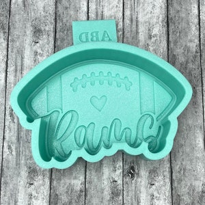 May include: A light green silicone mold shaped like a football with the word "Family" written in a cursive font and a small heart in the center. The mold has a raised design of a football with laces and stripes.