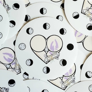 May include: A white sticker with a black outline of a heart shape. Inside the heart are two circles with a lavender flower bouquet in between. The circles are filled with a light beige color. The sticker is surrounded by black crescent moons with white stars.