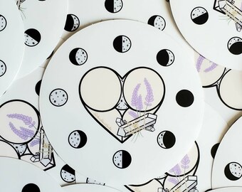 Puede incluir: Una pegatina blanca con un contorno negro en forma de corazón. Dentro del corazón hay dos círculos rellenos de un color beige claro, con un ramo de flores de lavanda entre ellos. La pegatina está rodeada de medias lunas negras con estrellas blancas.