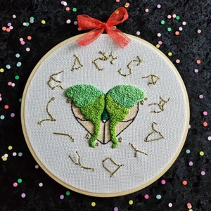 May include: A white embroidery hoop with a green butterfly design and gold stitching. The butterfly is in the center of the hoop and is surrounded by a circle of gold stars. The hoop is on a black background.