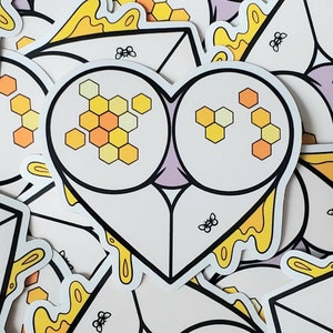 Può includere: Un adesivo bianco a forma di cuore con un design a nido d'ape e miele che cola. L'adesivo ha un contorno nero e una piccola ape in basso.