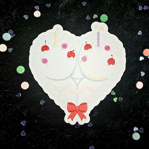 May include: A heart-shaped sticker with a cake design. The sticker features cherries, candles, and a red bow. The design is white with light blue accents and is set against a black background with confetti.