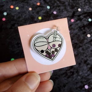 May include: A heart-shaped clear acrylic charm featuring a pink and white boba tea design. The boba tea has a straw and is filled with black and purple boba pearls.