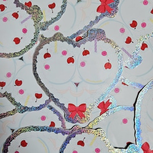 May include: A collection of heart-shaped stickers with a holographic, glittery border. Each sticker features a white background with red cherries, pink flowers, and a red bow. Some stickers have a purple candle.