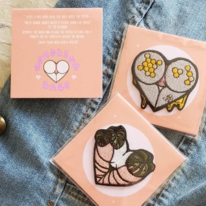 May include: Three iron-on patches on pink cards, featuring embroidered designs. One patch has a heart-shaped design with a honeycomb pattern, another with a plant, and the third with a butt design and the text "DEADBEAT BABE".