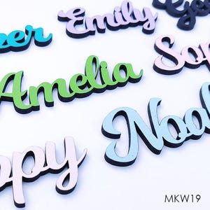 May include: A selection of wooden name cutouts in different colours and fonts. Names such as "Emily", "Amelia", and "Noel" are visible, each with a black outline. The names are painted in pink, green, and blue. The letters are approximately 7.6 cm long.