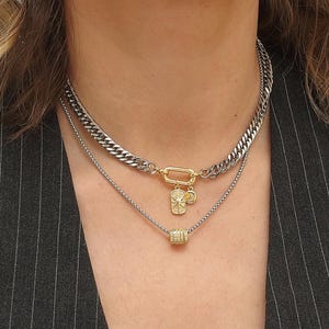 Può includere: Un set di collane a strati con una catena argentata spessa, una catena argentata delicata e una catena dorata con ciondoli. Le collane sono indossate sopra un gilet gessato grigio scuro. L'immagine include il testo "MonMaari 2025".