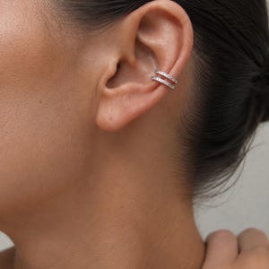 Può includere: Un ear cuff color argento caratterizzato da due fasce parallele decorate con piccole pietre chiare e scintillanti. Il gioiello è posizionato sulla cartilagine dell'orecchio, creando un aspetto stratificato grazie al design a doppia fascia.