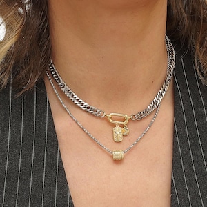 May include: A layered necklace set featuring a chunky silver chain, a delicate silver chain, and a gold-toned chain with a charm. The chains are worn over a black and white pinstripe vest.
