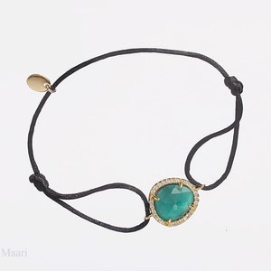 Può includere: Braccialetto con cordino nero con montatura color oro che tiene una grande gemma ovale color verde acqua, circondata da piccole pietre trasparenti. Un piccolo ciondolo color oro è attaccato al cordino.