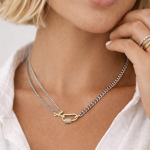 May include: A silver and gold-tone necklace featuring a double chain design. The necklace has a gold-coloured clasp with a row of small, clear stones. The person is wearing a white shirt. Several gold rings are on the person's finger.
