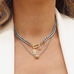 May include: A layered necklace set with a chunky silver chain and a finer silver chain with gold details. The top chain has a large clasp. The lower chain has a pendant and a small cylinder with rhinestones.