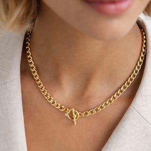 Könnte beinhalten: Eine goldfarbene Kette mit Knebelverschluss. Die Halskette ist im Stil einer klobigen Panzerkette gehalten, und der Verschluss hat ein kreisförmiges Design mit einem geraden Stangenverschluss. Die Halskette wird über einem hellen Blazer getragen.