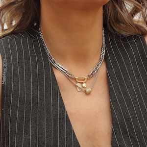 May include: A layered necklace with a silver chain and a gold-toned clasp. The necklace features a small pendant with a gold-toned charm and a small, round, gold-toned charm. The model is wearing a gray pinstripe vest.