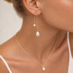 May include: A gold-coloured jewellery set comprising drop earrings and a necklace. The earrings feature a small pearl and a larger, teardrop-shaped pearl. The necklace has a matching teardrop pearl pendant. The jewellery is delicate and elegant.